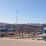 Bus terminal with several white and turquoise minibuses under sheltered bays; urban skyline in the distance.