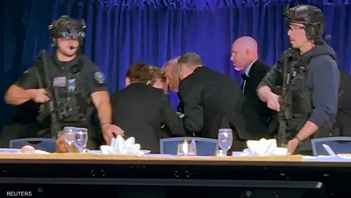 Security guards in tactical gear flank a conference table on a stage as suited people lean in for a discussion.