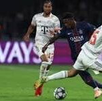 Two soccer players chase the ball on a green pitch during a match; white jersey player reaches for the ball as a purple-team opponent closes in.