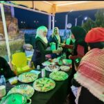 Group of people wearing head coverings around a table of food at an outdoor night event under a yellow canopy.