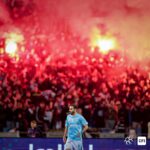 Soccer player in a light blue kit walks on the pitch as a red-flare crowd erupts in the background.