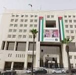 Facade of a large white multi-story office building with a wide entrance and a vertical green, white, and red stripe on the facade; cars parked along the street in front.