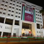 Night view of a white government building at night with a large banner featuring portraits and Arabic text, bordered by Jordanian flag colors and lights along the fence