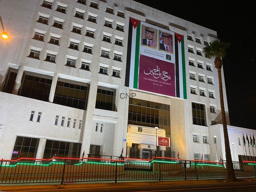 dam - Center News Network Night view of a white government building at night with a large banner featuring portraits and Arabic text, bordered by Jordanian flag colors and lights along the fence