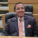 dayat - Center News Network Man in a dark suit with a peach tie sits at a desk in a formal courtroom or council chamber.