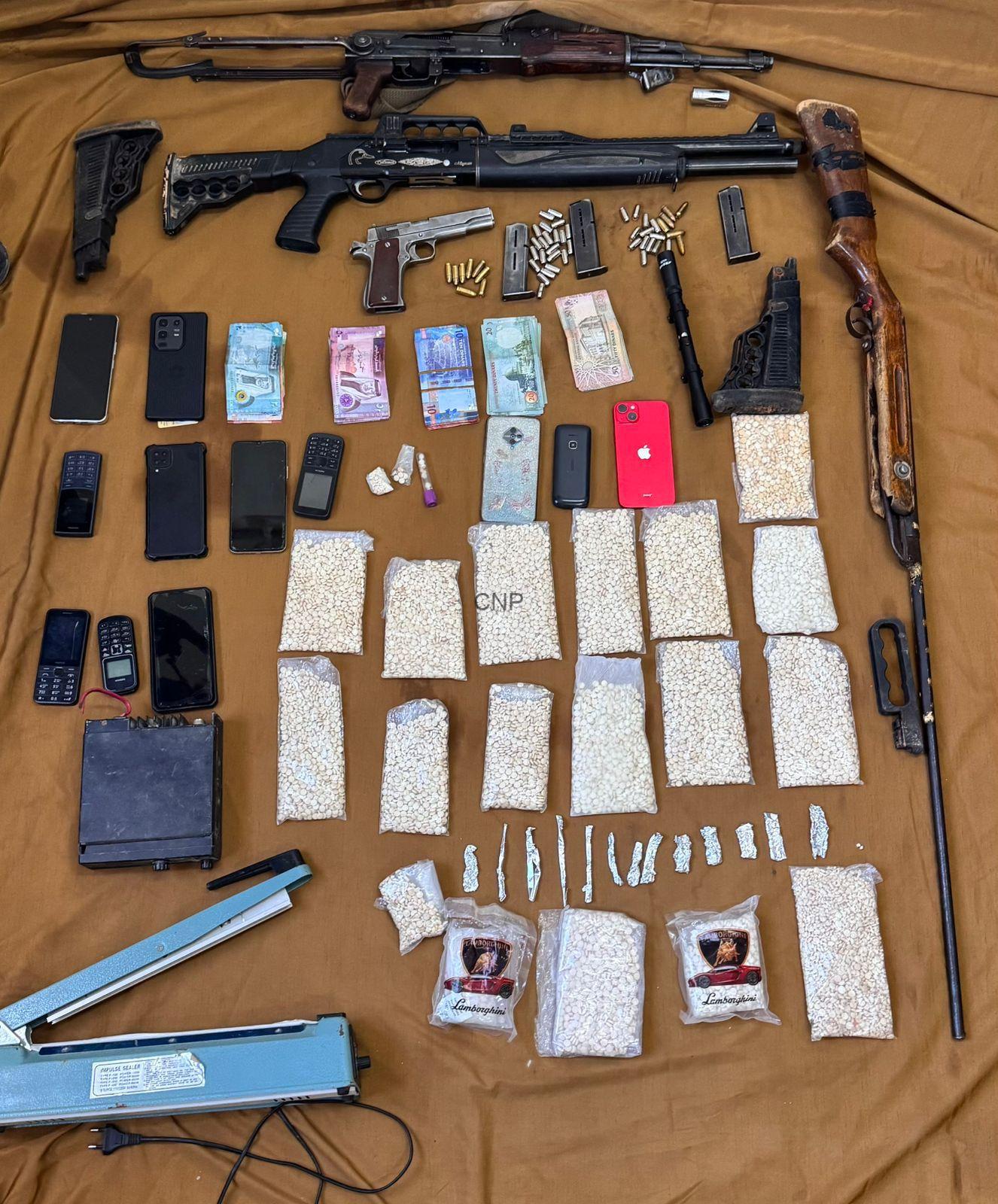 Weapon and contraband stash: several firearms, magazines, and ammunition laid out with money and phones on a brown backdrop for display or evidence.