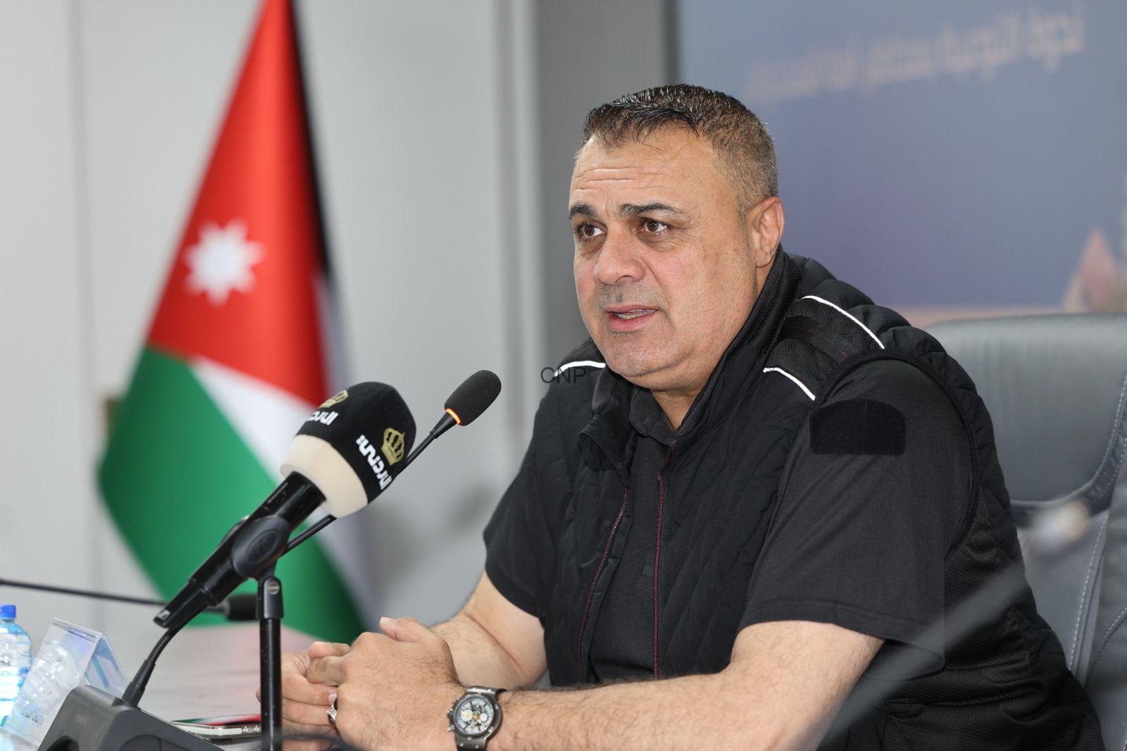 dru - Center News Network Man speaks at a press conference, seated at a table with a microphone in front, Jordan flag in the background.