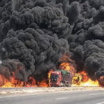 Two trucks on a highway with intense flames and thick black smoke rising behind them, indicating a large fire nearby.
