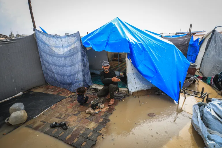 Man crouching at the entrance of a blue tent in a flooded refugee camp, a child nearby, with muddy ground and tarps around.