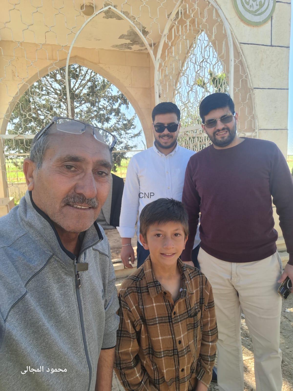 Four people pose for a selfie outdoors beside an ornate stone arch gate; foreground man in a gray jacket with glasses, a boy in a plaid shirt, and two men in sunglasses behind him.