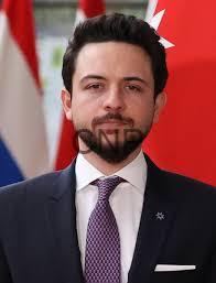 hai - Center News Network Professional portrait of a bearded man in a navy suit and purple tie, standing before red and white flags in the background.