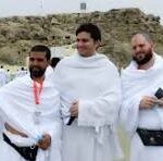 Three men in white robes pose together on a sandy beach during a ceremony, with onlookers and distant rocks in the background.