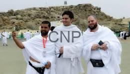 hhh - Center News Network Three men in white robes pose together on a sandy beach during a ceremony, with onlookers and distant rocks in the background.
