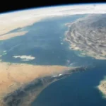 From space, a curved coastline with blue sea between arid desert to the left and rugged land to the right, showing Earth’s horizon rim and cloud-free atmosphere.