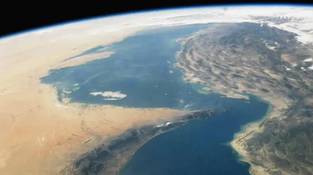 From space, a curved coastline with blue sea between arid desert to the left and rugged land to the right, showing Earth’s horizon rim and cloud-free atmosphere.