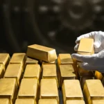 Gloved hands stack gold bars in a vault, with one bar being placed on top of the stack.