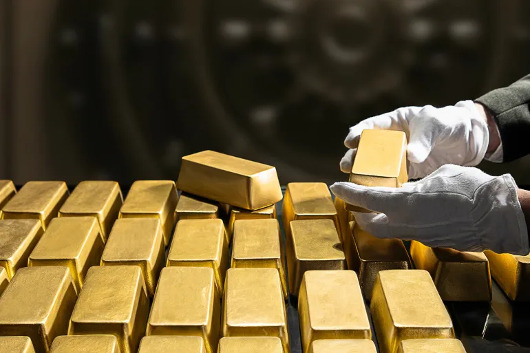 Gloved hands stack gold bars in a vault, with one bar being placed on top of the stack.