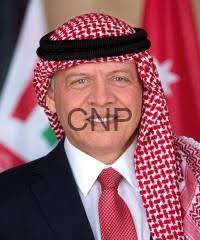 Portrait of a man wearing a red-and-white keffiyeh and dark suit, with flags blurred in the background.