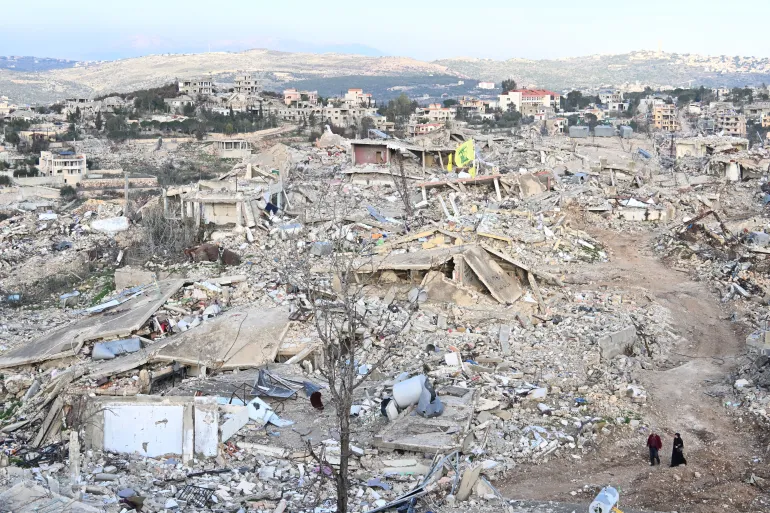 lob - Center News Network Rubble-strewn hillside town with collapsed buildings and debris, two people walking along a dirt path in the foreground.