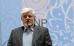 Older man with white hair and glasses in a dark suit speaks at a podium during a conference, with a blue patterned backdrop.