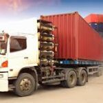 White semi-truck hauling stacked shipping containers on a flatbed in a busy container yard. [] Containers in red, blue, and orange are visible in the background.