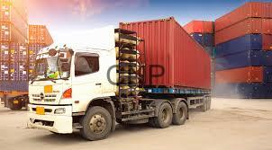 White semi-truck hauling stacked shipping containers on a flatbed in a busy container yard. [] Containers in red, blue, and orange are visible in the background.