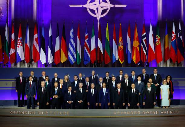 Group photo of world leaders at a NATO summit, with numerous national flags and the NATO emblem above them on a purple-lit backdrop.