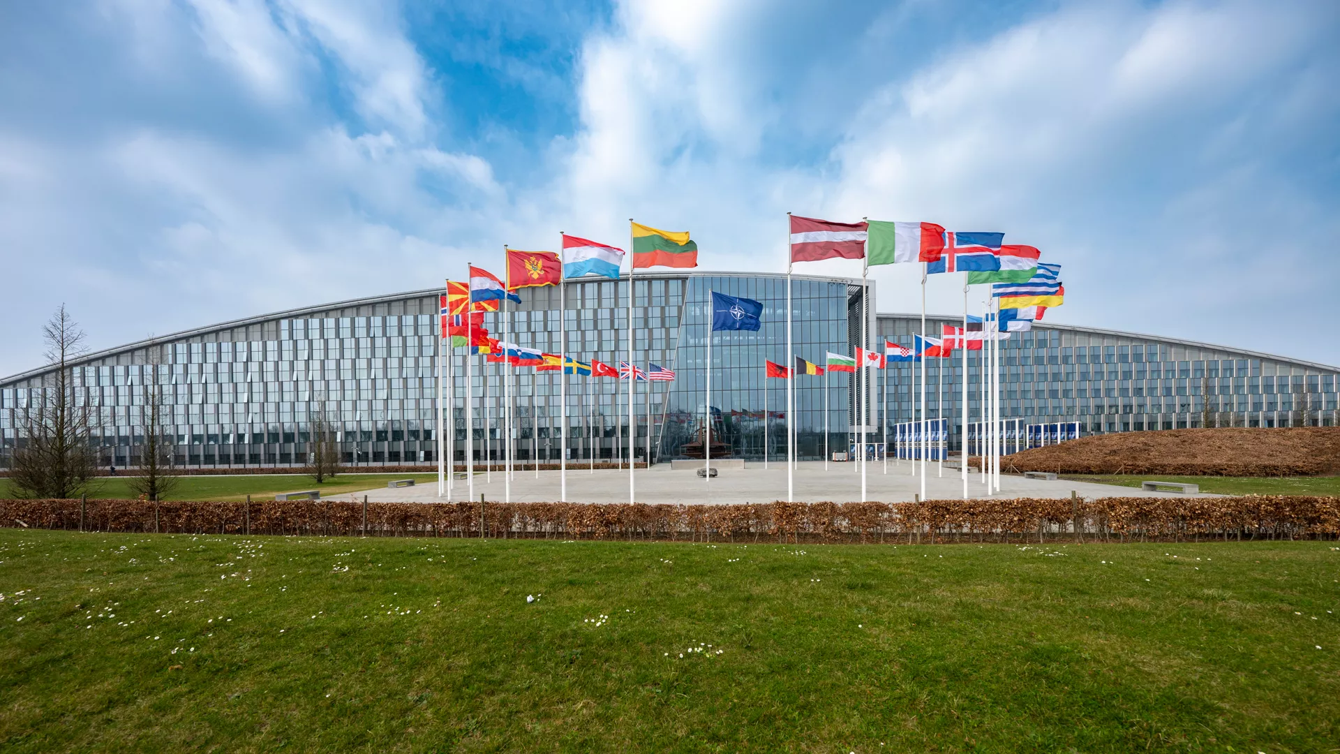 nato - Center News Network Glass-fronted modern building with a courtyard and a row of international flags on tall poles in front, blue sky above.