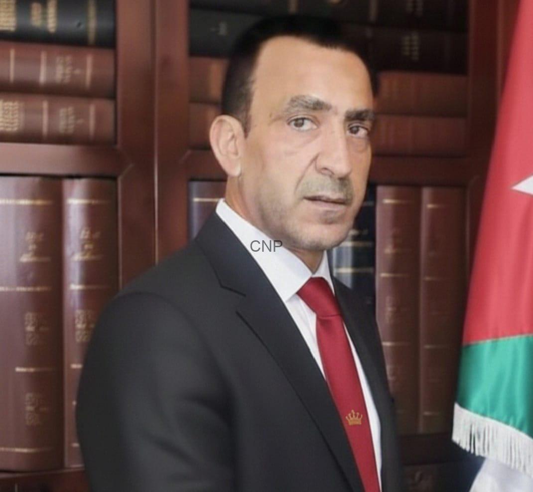 Man in a dark suit and red tie standing in front of a bookshelf with leather-bound books and a Jordanian flag nearby.