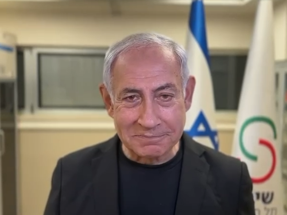 Older man in a dark blazer speaks to the camera, with flags in the background.