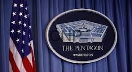 Seal of the Pentagon on a blue curtain backdrop with the U.S. flag to the left.