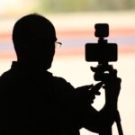 Silhouette of a photographer adjusting a camera on a tripod at sunset.