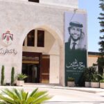 Stone building with an arched entrance and a large vertical banner depicting a man in traditional Arab dress; banner includes English text 'Celebrating Al Hussein' and Arabic script, surrounded by potted plants and trees at the entrance.