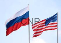 Russia and United States flags waving on flagpoles against a pale blue sky.