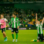 Soccer players in green-and-white uniforms celebrate on a bright stadium field, with one kneeling and pointing to the sky as teammates smile nearby.