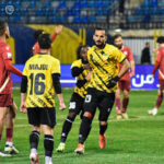 Bearded player in yellow and black kit dribbles the ball during a match, with red-clad opponents nearby on a green field at the stadium.