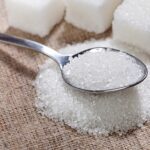 Spoon scooping white granulated sugar on a burlap surface with sugar cubes in the background.