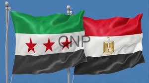 Two national flags waving against a blue sky: Egypt on the right and a green‑white‑black flag with three red stars on the left, both on flagpoles.