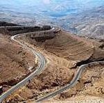 Switchback road zigzags down a rocky desert canyon with brown hills and distant mountains in the background.