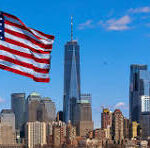 American flag waving above the Manhattan skyline with tall buildings against a blue sky, sunny day.]?