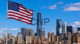 American flag waving above the Manhattan skyline with tall buildings against a blue sky, sunny day.]?