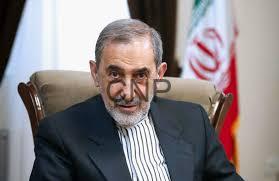 Older man with gray hair and beard in a dark suit seated in an office, with an Iranian flag in the background.