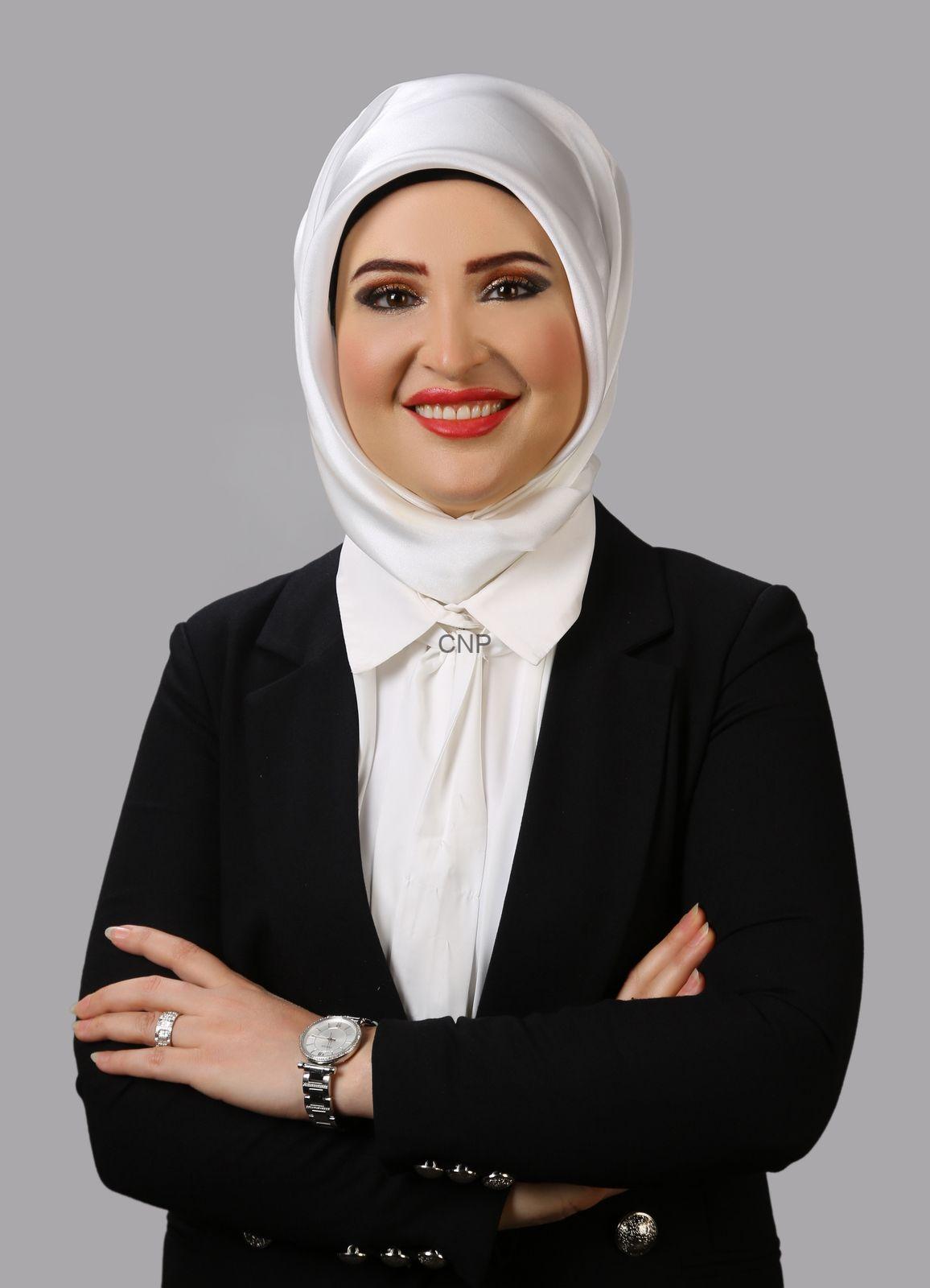 Professional portrait of a smiling woman wearing a white hijab, white blouse, and black blazer with arms crossed against a gray background.