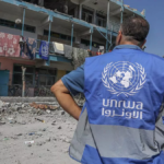 Back of a UNRWA worker in a blue vest surveying rubble and damaged buildings in a destroyed street.