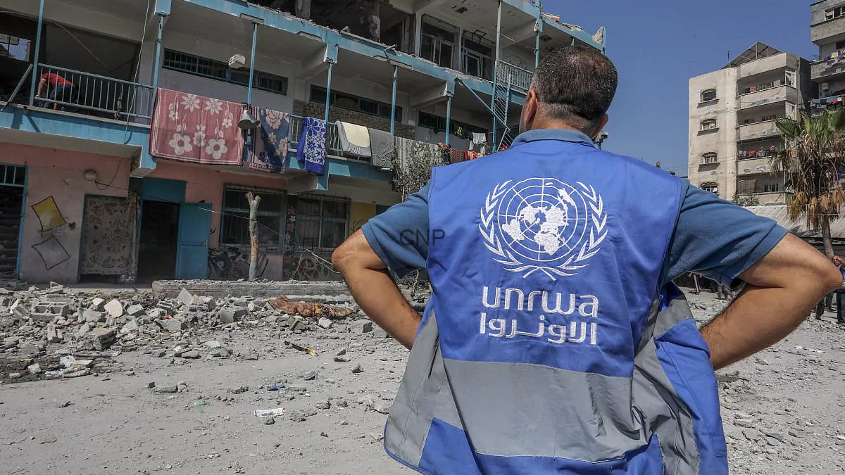 04d2ba93-e969-4746-958e-3cbeb3e7d7f8_16x9_1200x676 - Center News Network Back of a UNRWA worker in a blue vest surveying rubble and damaged buildings in a destroyed street.