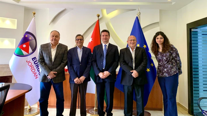 Five businesspeople in suits stand in a diplomatic office with Jordan, Turkey, and EU flags behind them.