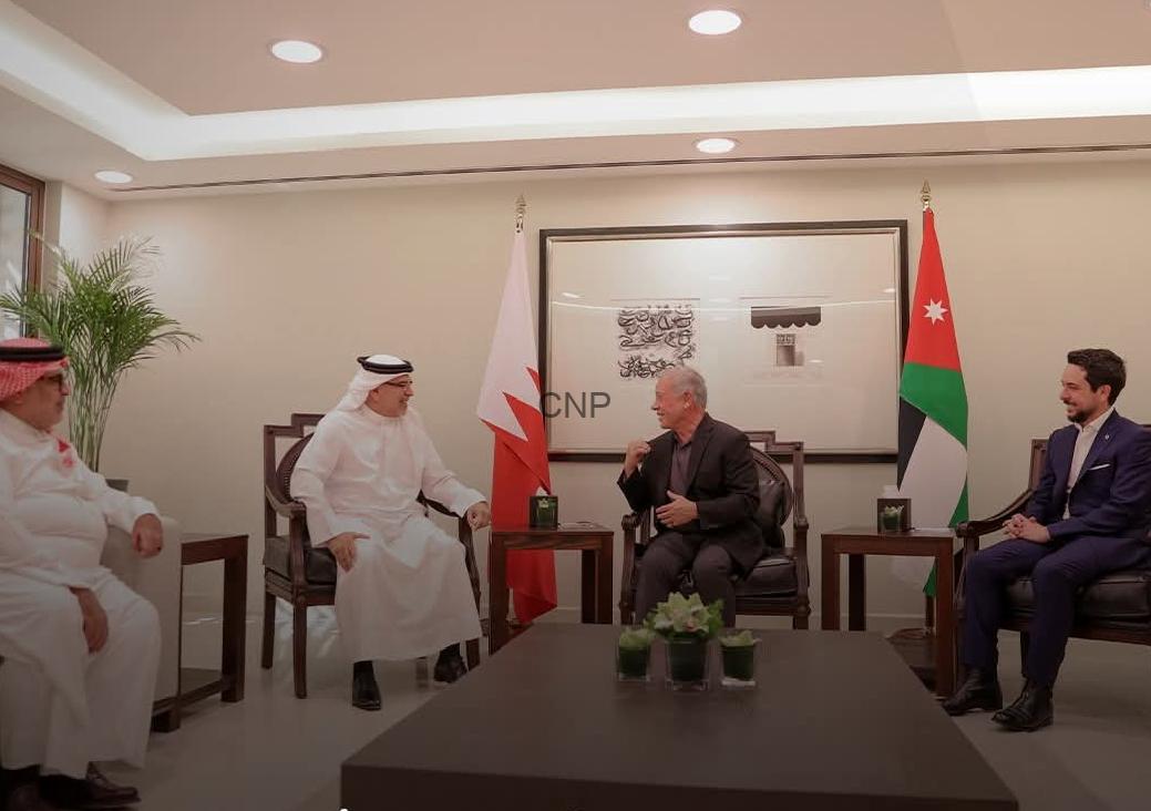 Diplomatic meeting in a formal room with four men seated, flags behind them and a central coffee table with plants.