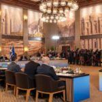 Delegates seated around a U-shaped conference table in a grand hall with murals, chandeliers, and press photographers at the back.
