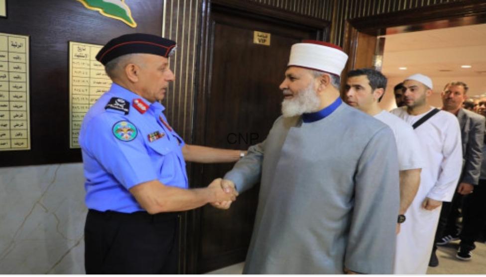 Police officer in a blue uniform shakes hands with a bearded man in a gray traditional robe and white cap as others stand in line nearby.