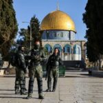 Group of soldiers in tactical gear standing in a stone courtyard with the Golden Dome of the Rock in Jerusalem in the background.